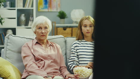 cute little teen girl and her grandmother sitting on the sofa at home and watching something scary on tv, then granddaughter hiding her eyes and being afraid