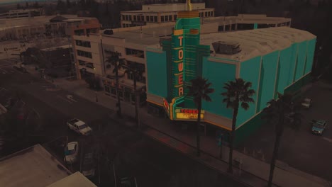 Cinematic-top-city-aerial-view-of-downtown-Roseville,-California-with-industrial-buildings,-shops-and-tower-theater-next-to-a-multi-level-parking-lot-structure-during-sunset-and-cars-passing-by