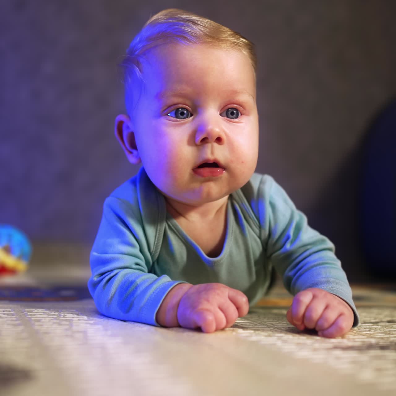 Liten babygutt med lyst hår og store øyne ser interessert rundt seg. Spedbarnet ligger på gulvet hjemme.