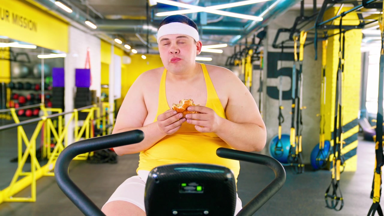 ジムでエアロバイクに座りながらハンバーガーを食べる太りすぎの男性