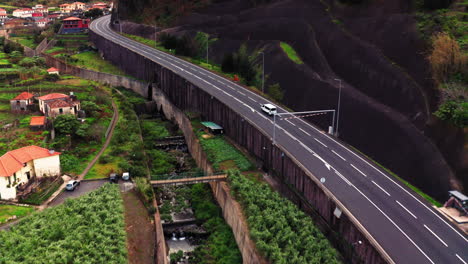 Luftaufnahme-Eines-Lieferwagens,-Der-Auf-Einer-Asphaltierten-Straße-Auf-Der-Insel-Madeira-Fährt,-Umgeben-Von-Grüner-Landschaft,-Bergen-Und-Alten-Gebäuden