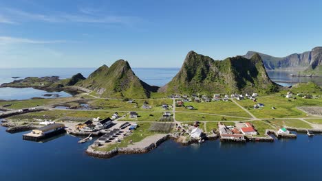 Aerial view of Værøy harbor: colorful village by the sea, dramatic mountains rising behind, and the endless blue of the Norwegian coast