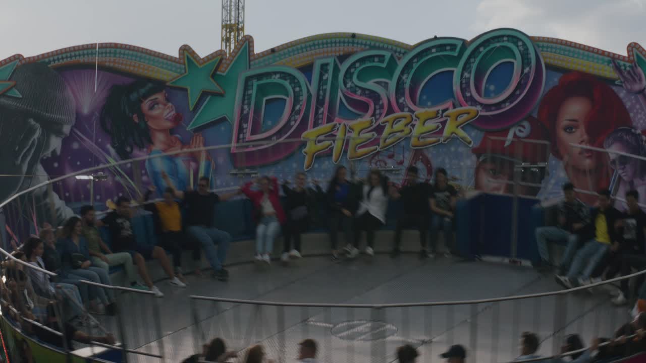 People walking by as others are riding a spinning disco wheel at Balvarian festival at Stuttgart, Germany, Europe at sunset, panning view angle