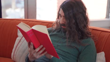 Hispanic male with long hair and beard happily sitting on orange couch reading from red book