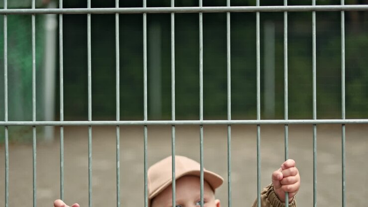 Un ragazzo fa capolino da dietro una grata di metallo per strada. Il bambino gioca all'aperto ed esplora l'ambiente. Il ragazzo gioca al parco giochi