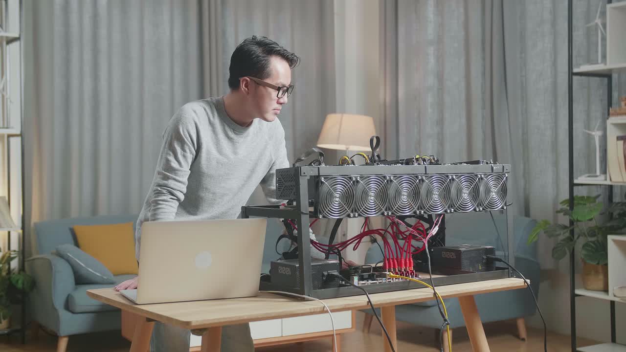 안경을 쓴 아시아인 남자가 노트북으로 일하면서 암호화폐 채굴 기계를 보고 있다