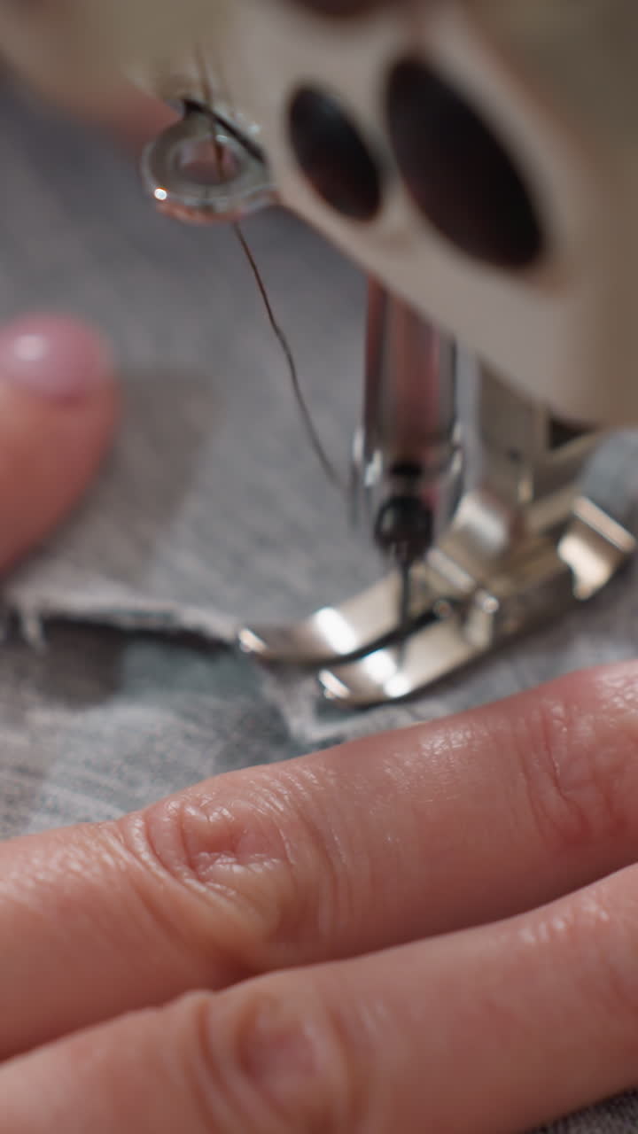 Primer plano extremo de dedos caucásicos cerca de una aguja de coser que guía el borde de una tela gris, pie prensatelas y dientes de arrastre, uñas pulidas cerca del hilo y la bobina, acción de dobladillo cuidadosa, luz de estudio suave, enfoque