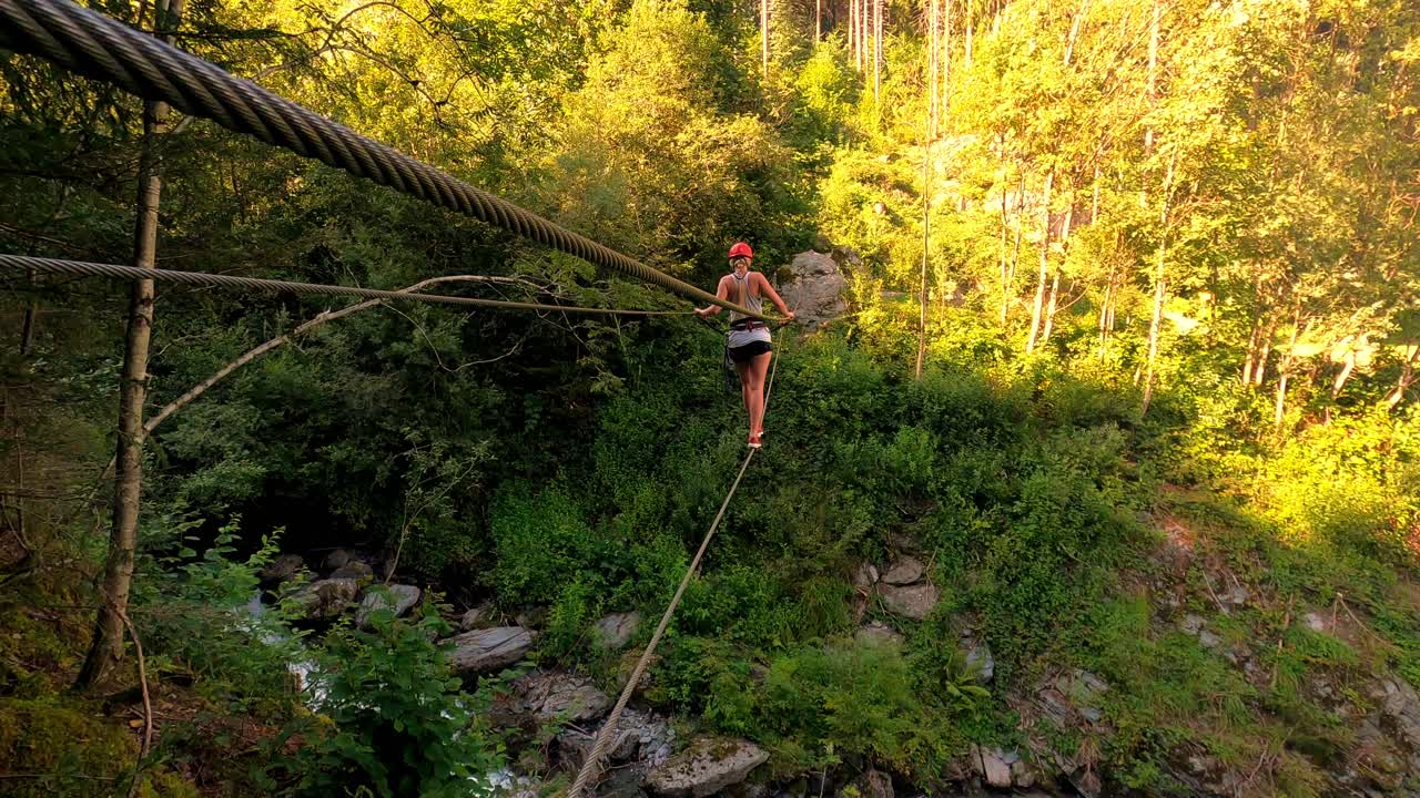 검은색 핫팬츠, 흰색 티셔츠, 빨간색 헬멧을 쓴 금발 소녀가 흔들리는 케이블 다리를 건너고 있습니다.