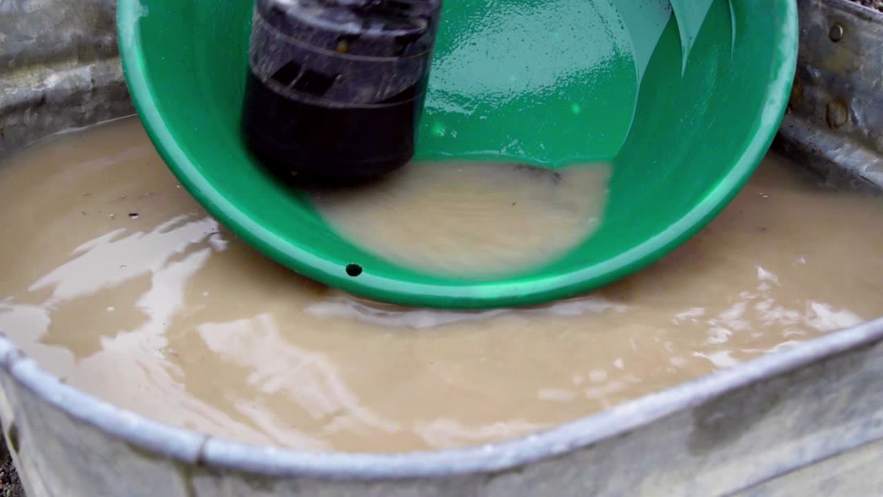 Panning for gold in green trap pan with black magnet to remove grunge, silt, sand, and clay alluvial deposits and washing in tin bucket of brown murky water, static close up portrait