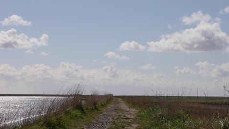 Fußweg,-Wasser-Auf-Der-Linken-Seite,-Sumpfgebiet-Auf-Der-Rechten-Seite,-Horizont-Und-Skyline-Mit-Flauschigen-Wolken-Und-Sanft-Blauem-Himmel