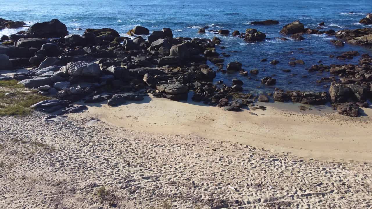 Dynamic drone footage transitioning from a near-cenital view of the sandy shoreline and rocks to a forward-facing perspective, flying smoothly over the sea, revealing an endless ocean horizon.