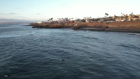 Aerial-view-of-surfers-on-surfing-boards-in-water-of-Pacific-Ocean-by-San-Diego-Cliffs-at-sunset,-drone-shot,-California-USA