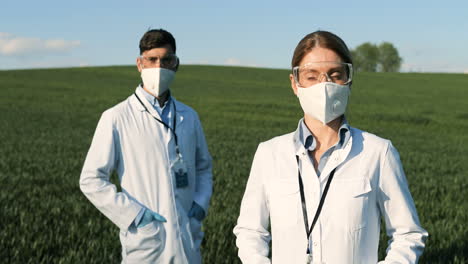 investigadores caucásicos con bata blanca, máscara y gafas mirando la cámara en el campo verde