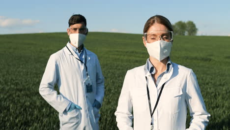 caucasian researchers in white gown, mask and googles looking at camera in the green field