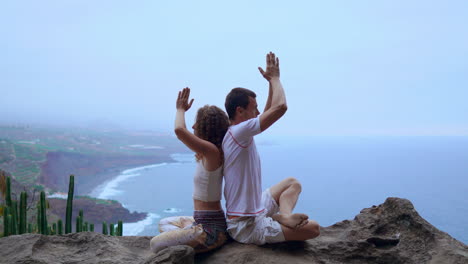 amidst mountain views and the ocean's horizon, a man and woman sit on a rock back to back, immersed in meditation and yoga