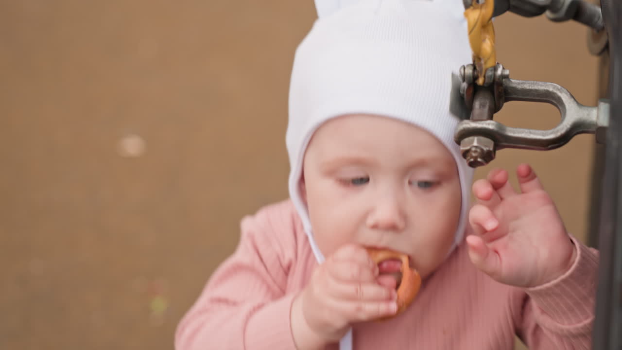 Baby Enjoys Treat Beside Playground Equipment, Young Child Tastes Snack Near Outdoor Climbing Apparatus, Curious Infant In Pink Romper Samples Snack Next To Swing And Metal Fixture