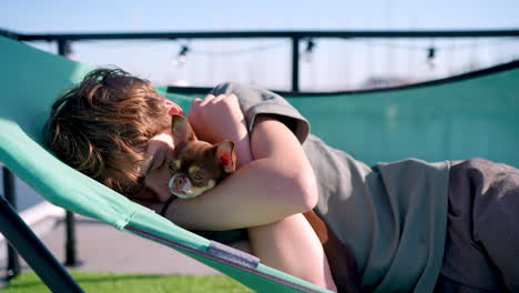 young boy peacefully resting on rooftop deck chair, tenderly hugging small chihuahua while enjoying warm sunlight and urban skyline backdrop