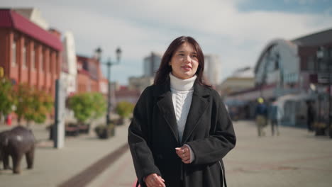 confident woman in black coat walking outdoors, adjusting her hair with a warm smile, surrounded by blurred urban background with people and buildings in the distance