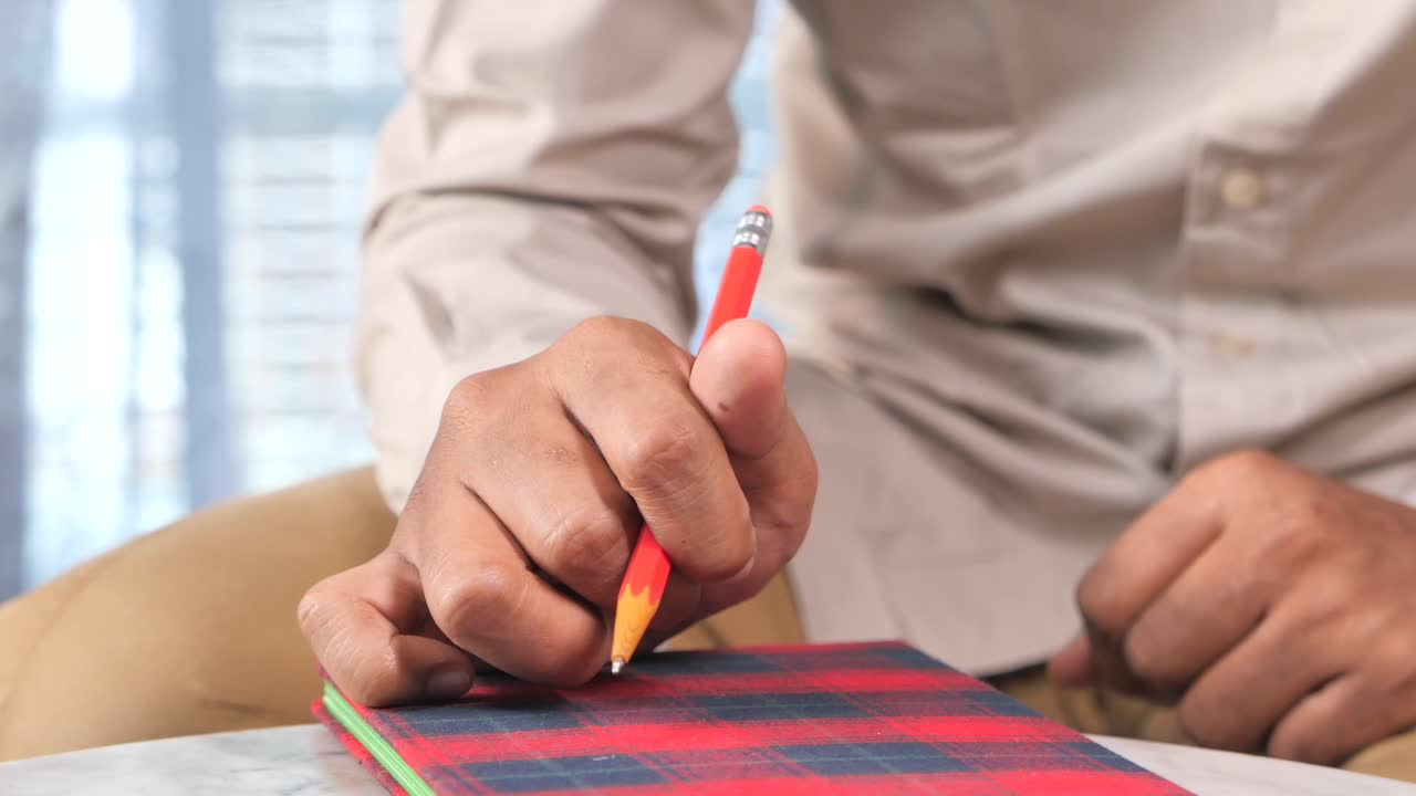 primer plano de la mano de un hombre escribiendo en un cuaderno con un lápiz rojo