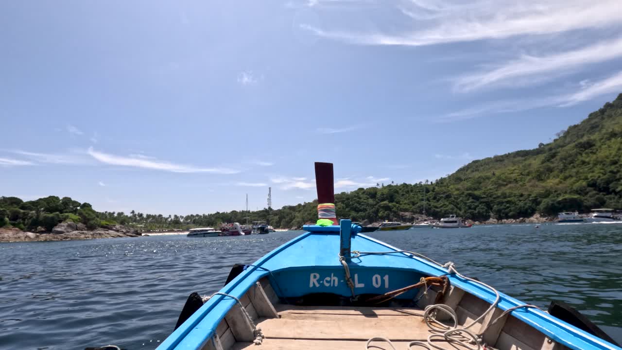 Wooden long-tail boat moves toward Phuket coastline, clear sky, midday sunlight, steady forward camera