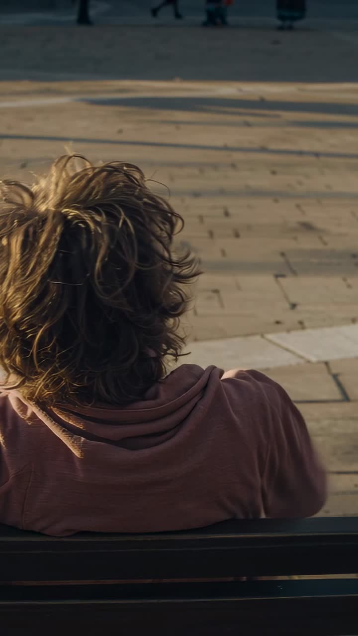 Vertical video: Turning man in pink hoodie leaning on wood bench in plaza, breeze blowing, relaxing