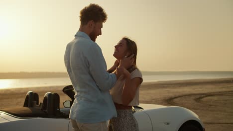 a happy blonde girl in a white top and her bearded boyfriend in a blue shirt are stroking each other's faces and hugging near a white convertible car. a romantic summer meeting against the river and the yellow sky