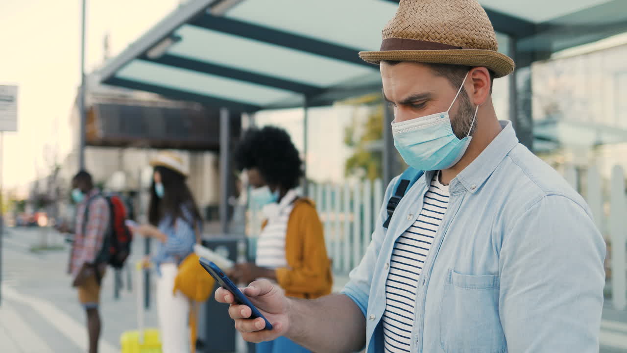 Caucasian man traveller in facial mask and hat using smartphone at bus stop while others travellers waiting for transport