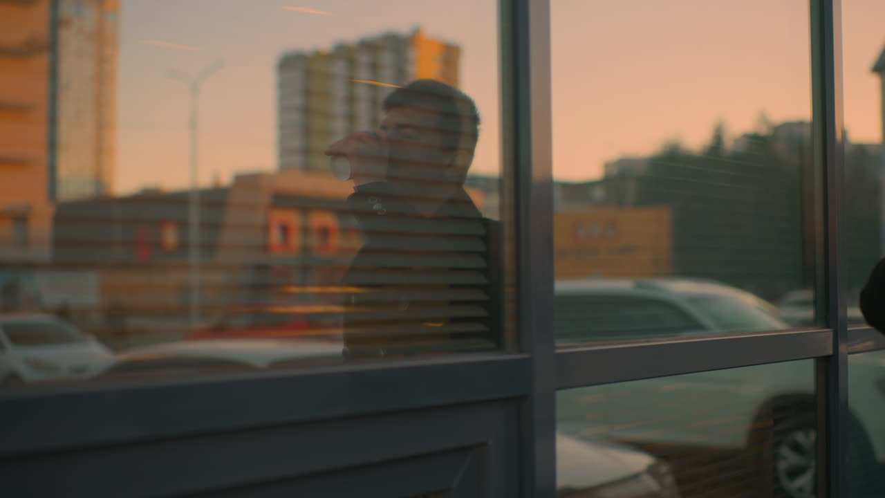 Close-up of glass reflecting red cloud, building, with person in black clothing walking while drinking coffee, people walking in urban environment, busy city street and parked cars