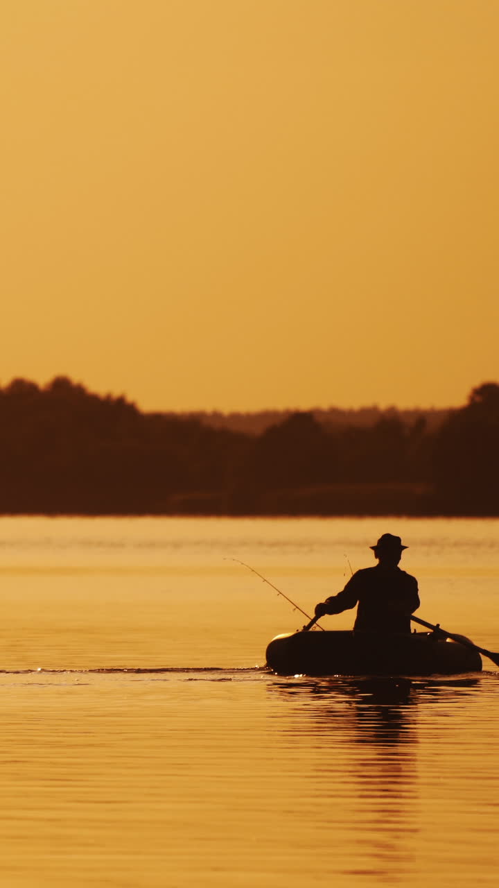 Silhouette of man sailing with two wooden oars on a small boat at sunset time. Beautiful sunset in the river and the silhouette of a man who tries to find a place for fishing on rowboat. Vertical video