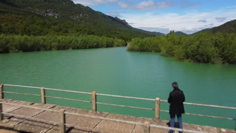 Aerial-view-of-a-man-standing-in-the-bridge-flying-backwards,-over-the-green-lake