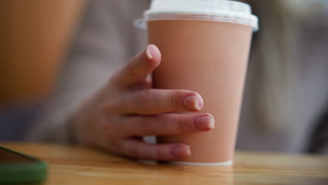 close-up hand view of lady tapping coffee cup on wooden table with phone beside it, background slightly blurred, capturing an intimate, calm moment in a cozy cafe setting