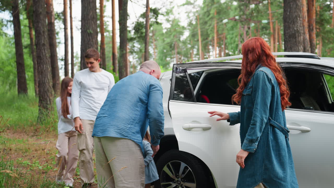 Family enjoys picnic moment in arboretum as father lifts youngest child from white car while mother watches warmly, surrounded by tall trees, serene summer outing full of family tenderness