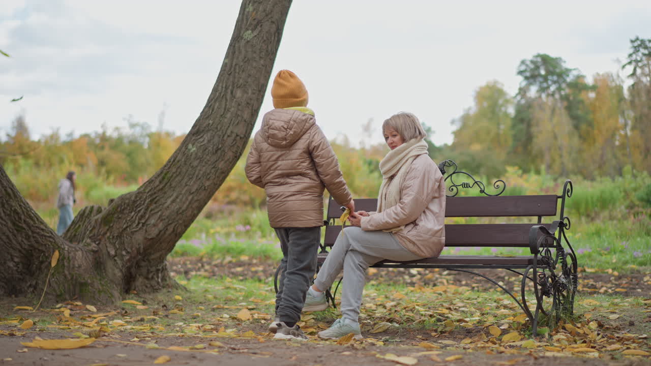 child walks up to mum focused on phone on bench in autumn park then gently pushes mum playfully causing laughter, wearing pink coat and chunky scarf amid golden leaves and blurred foliage