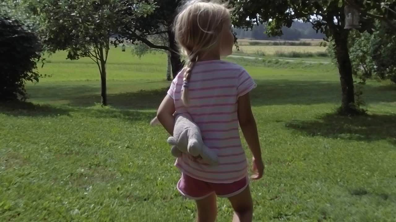 siguiendo a una linda niña de tres años con un animal de peluche caminando por un campo de hierba con manzanos en verano, cámara lenta