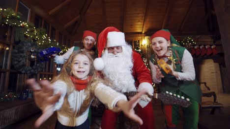 happy santa, elves and girl with musical instruments dancing and looking to camera. funny christmas atmosphere in santa's residence.