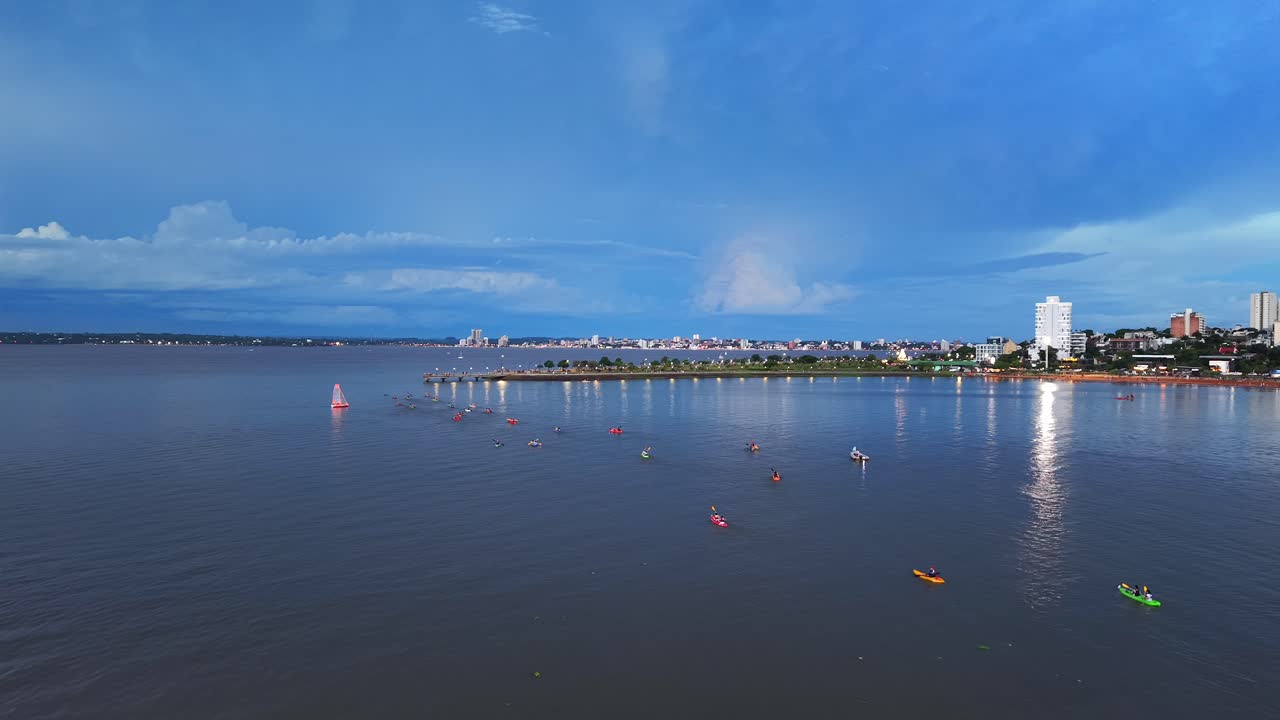 Filmato aereo di un folto gruppo di kayakisti che pagaiano insieme sul fiume Paraná. La scena cattura l'acqua calma e le luci della città al crepuscolo.