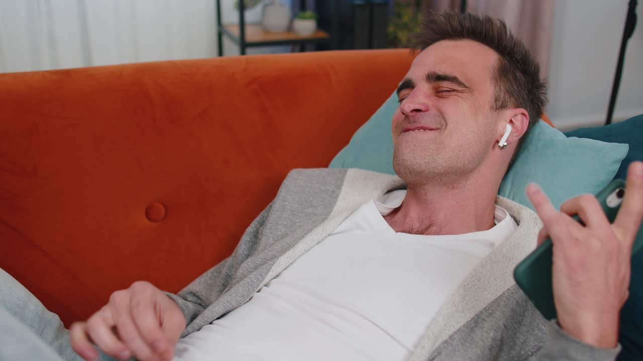 hombre feliz y relajado con auriculares inalámbricos bailando, escuchando su música favorita, acostado en el sofá en casa