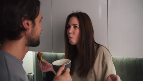 una feliz chica morena con una camiseta beige está tratando de conseguir un pedazo de pan atrapado en sus dientes y su novio moreno con paja la ayuda y se están divirtiendo juntos mientras pasan tiempo juntos por la noche en una cocina moderna
