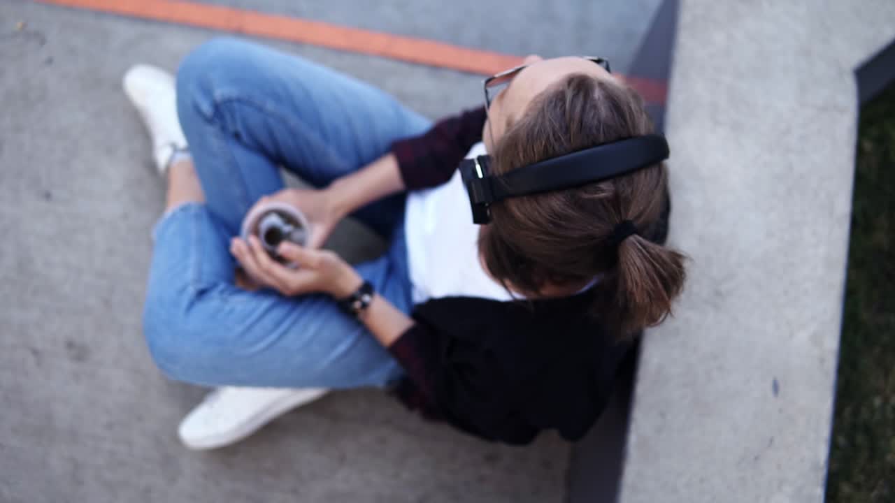 mujer joven relajada sentada en el suelo al aire libre y escuchando la música. usando vaqueros azules, camiseta blanca y abrigo negro