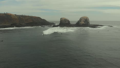 Aerial-of-Surfer-Riding-wave-next-to-giant-rocks-on-a-dark-cold-day-in-Pichilemu,-Chile-4K