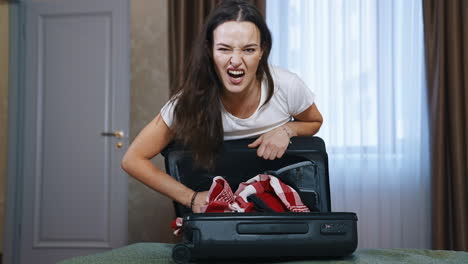 angry woman with suitcase indoors. young female trying to close the travel bag full of clothes in the hotel room. tourist girl packaging luggage.