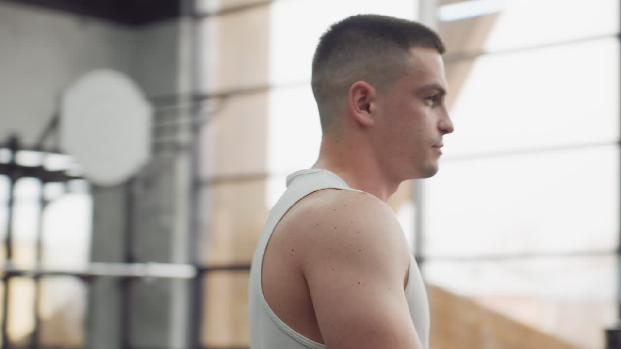 Young man with focused expression walks toward gym machine in modern fitness center with blurred exercisers under bright natural light and industrial steel beams interior design