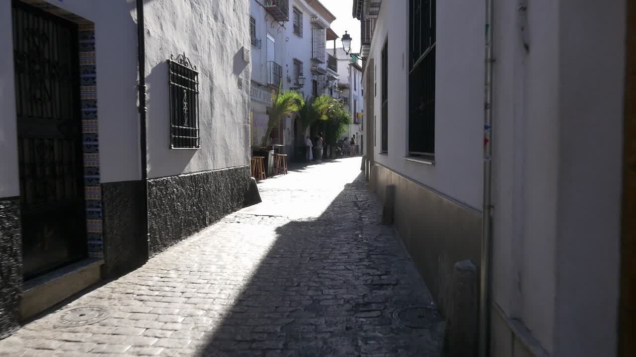 Vista in prima persona di qualcuno che cammina attraverso uno stretto e vuoto vicolo nell'Albayzín, con un forte contrasto di luci e ombre sulle pareti bianche e sul pavimento in ciottoli. Città mediterranee tradizionali.