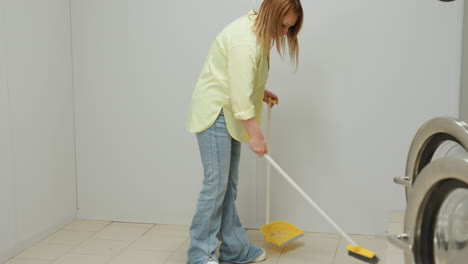 Downward view shows business owner in yellow shirt, jeans, white sneakers sweeping tiled floor with broom and dust pan near industrial washers inside bright laundromat, routine cleaning