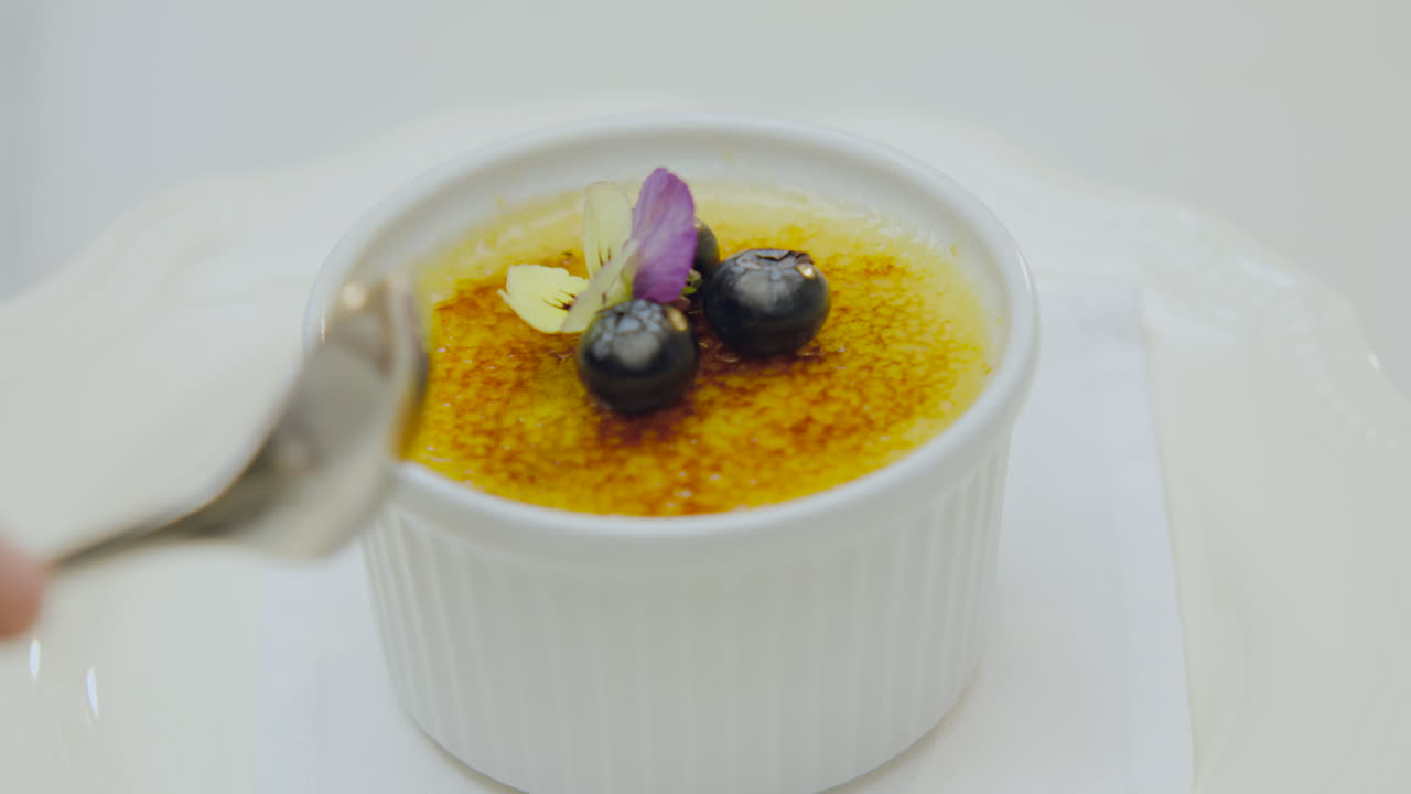 Crispy creme brulee dessert adorned with blueberries and a flower while a hand takes a portion with a spoon