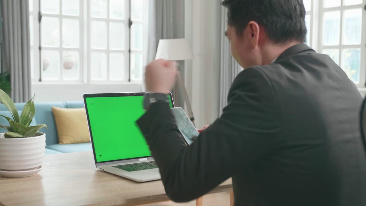 Asian Businessman Wearing Business Suit With Green Screen Computer Looking At Mobile Phone And Being Happy With Project Bid Result While Working At Home.