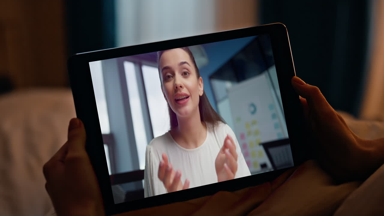 Freelancer che ascolta un coach durante una videoconferenza su tablet in una stanza serale, primo piano