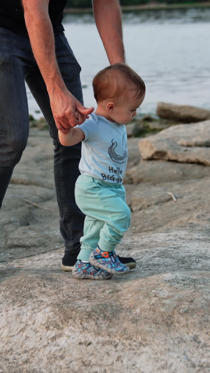 Little cute toddler stepping by the stones supported by his father. Loving dad teaching baby to walk in nature. River at backdrop. Vertical video