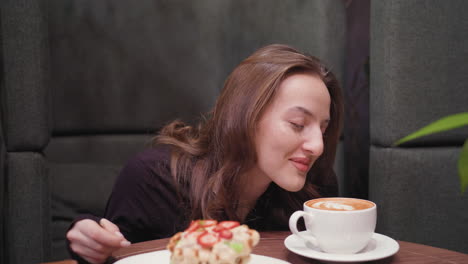 young woman with closed eyes and gentle smile leans toward latte on wooden table, savoring aroma of hot drink beside plate of waffles topped with strawberries in cozy room with soft lighting