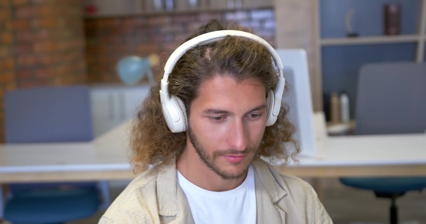 Man wearing white-headset hearing audio, leaning forward, nodding and reading laptop at office desk